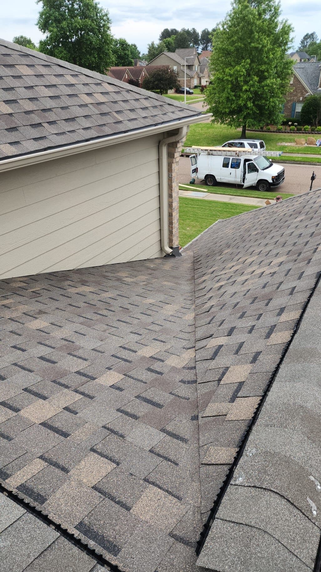 Edge of a freshly installed shingle roof with a work truck parked in the driveway below