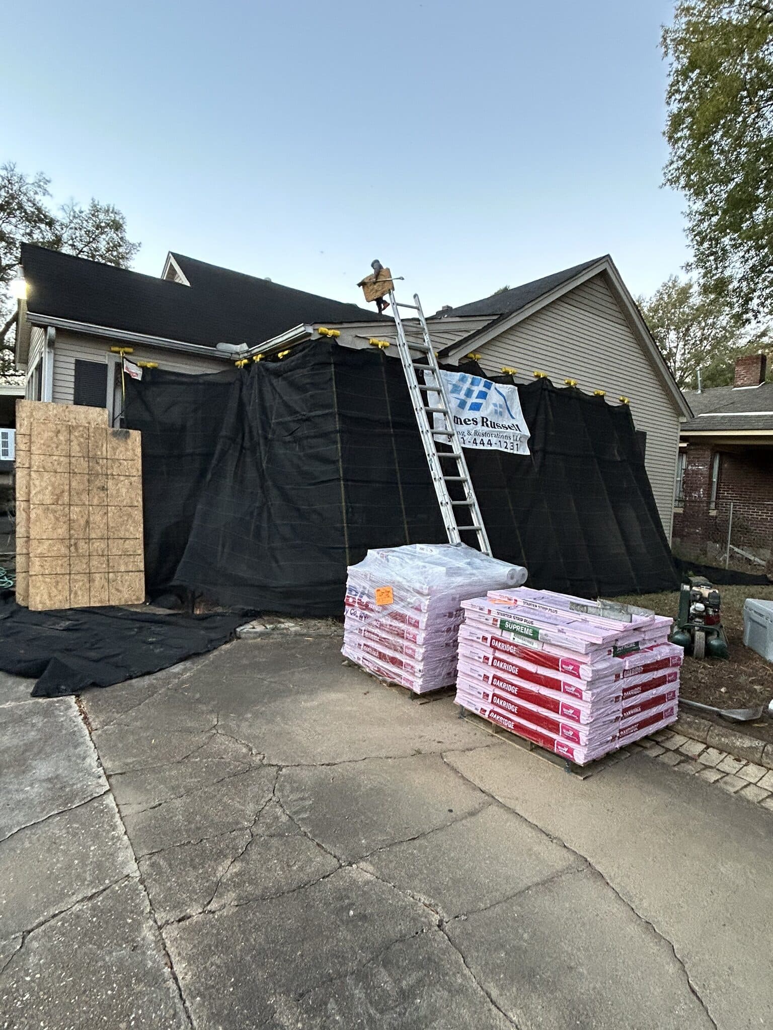 Pallets of shingle bundles staged in a driveway with a ladder to the tarped roof